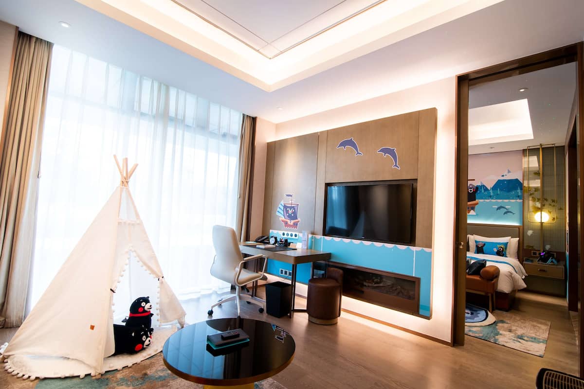 A modern hotel room with a large TV, a desk with a chair, a small tent, and a bed with a nightstand.