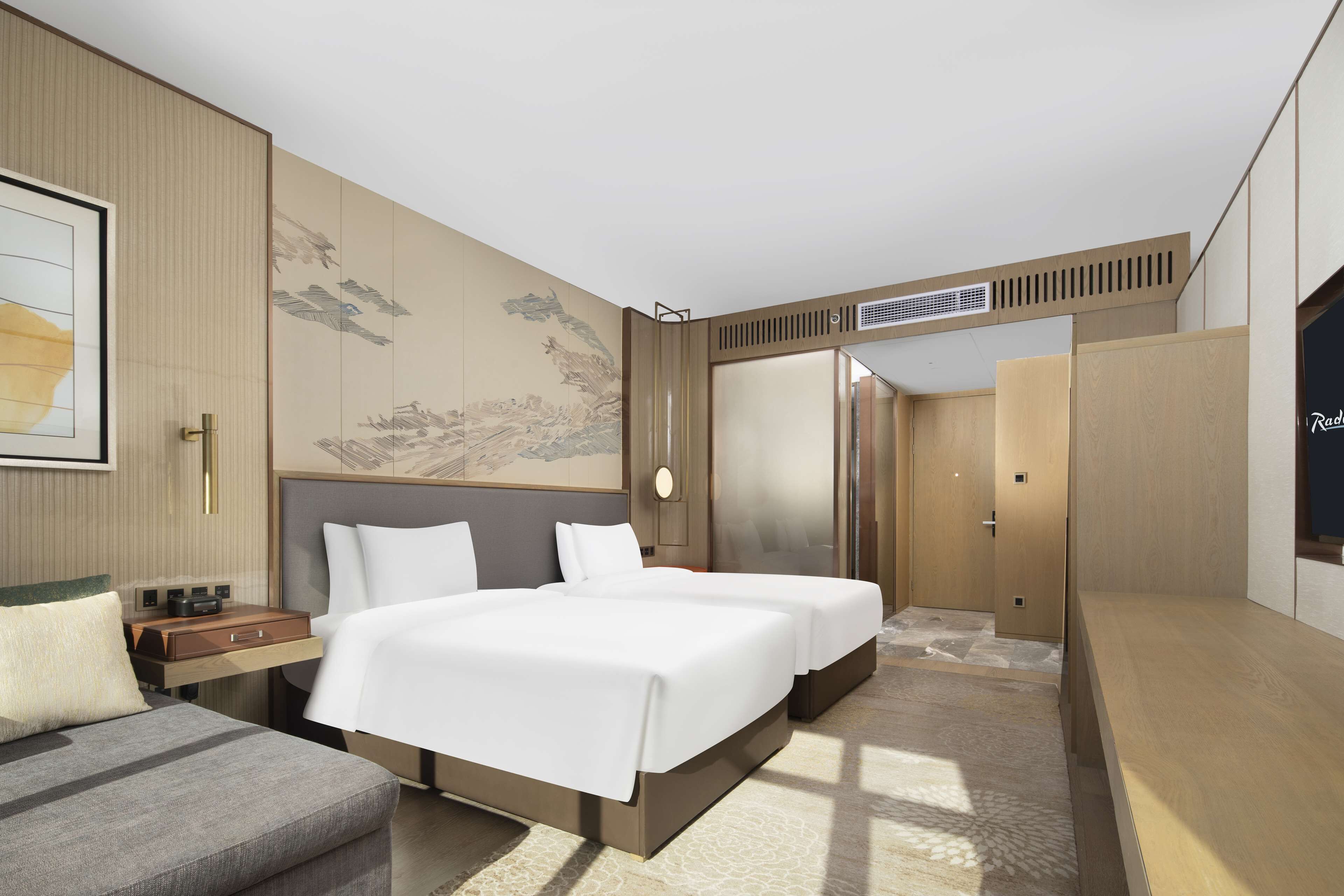 A modern hotel room with a large bed, a bedside table, a desk, and a wardrobe.