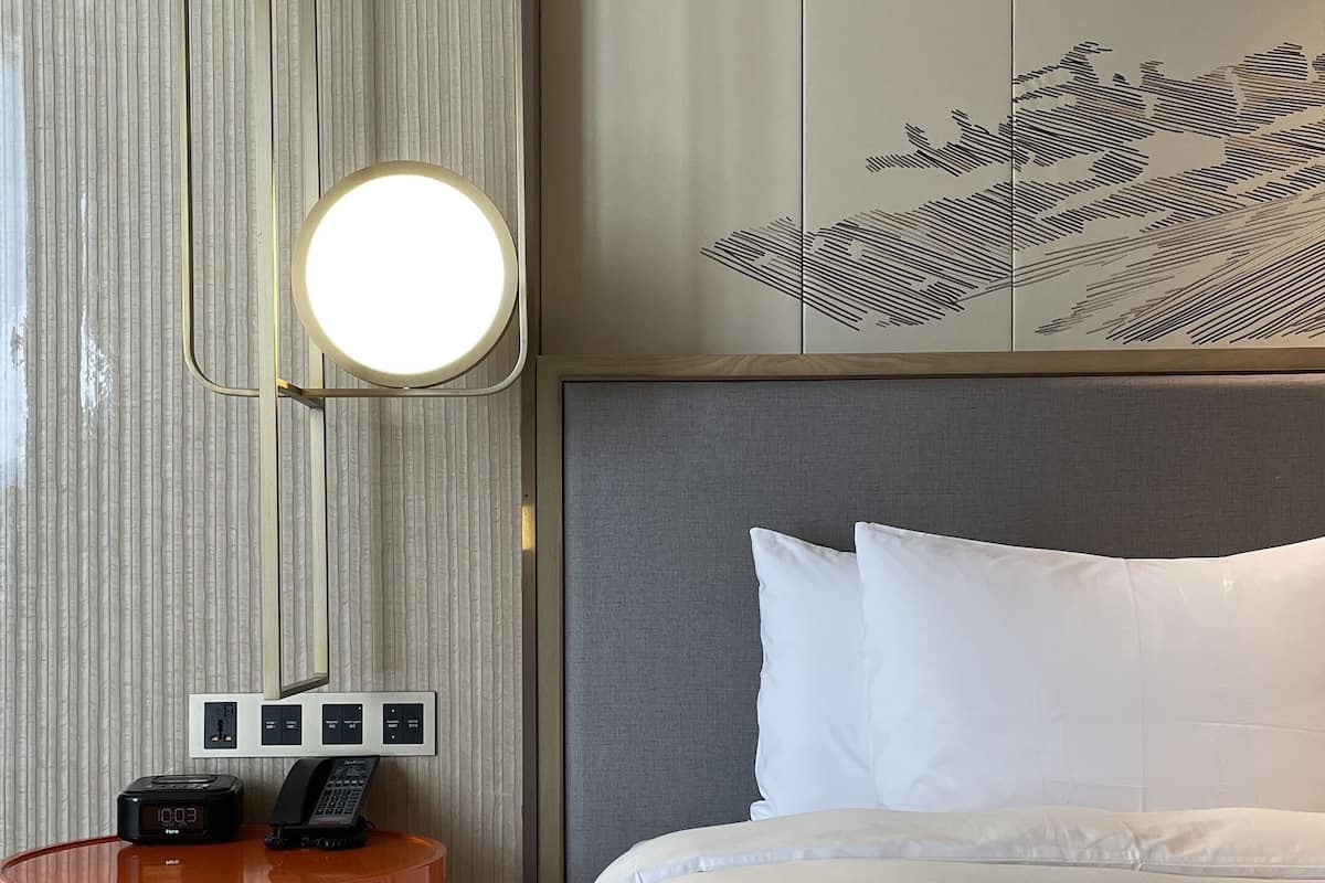 A hotel room with a bed, a round orange table with a lamp, and a wall with a decorative panel.
