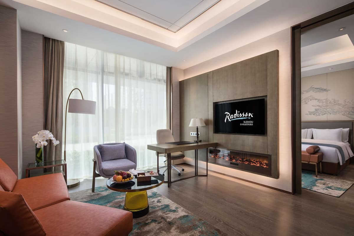 A modern hotel room with a flat-screen TV, a sofa, a chair, a small table with fruit, and a fireplace.