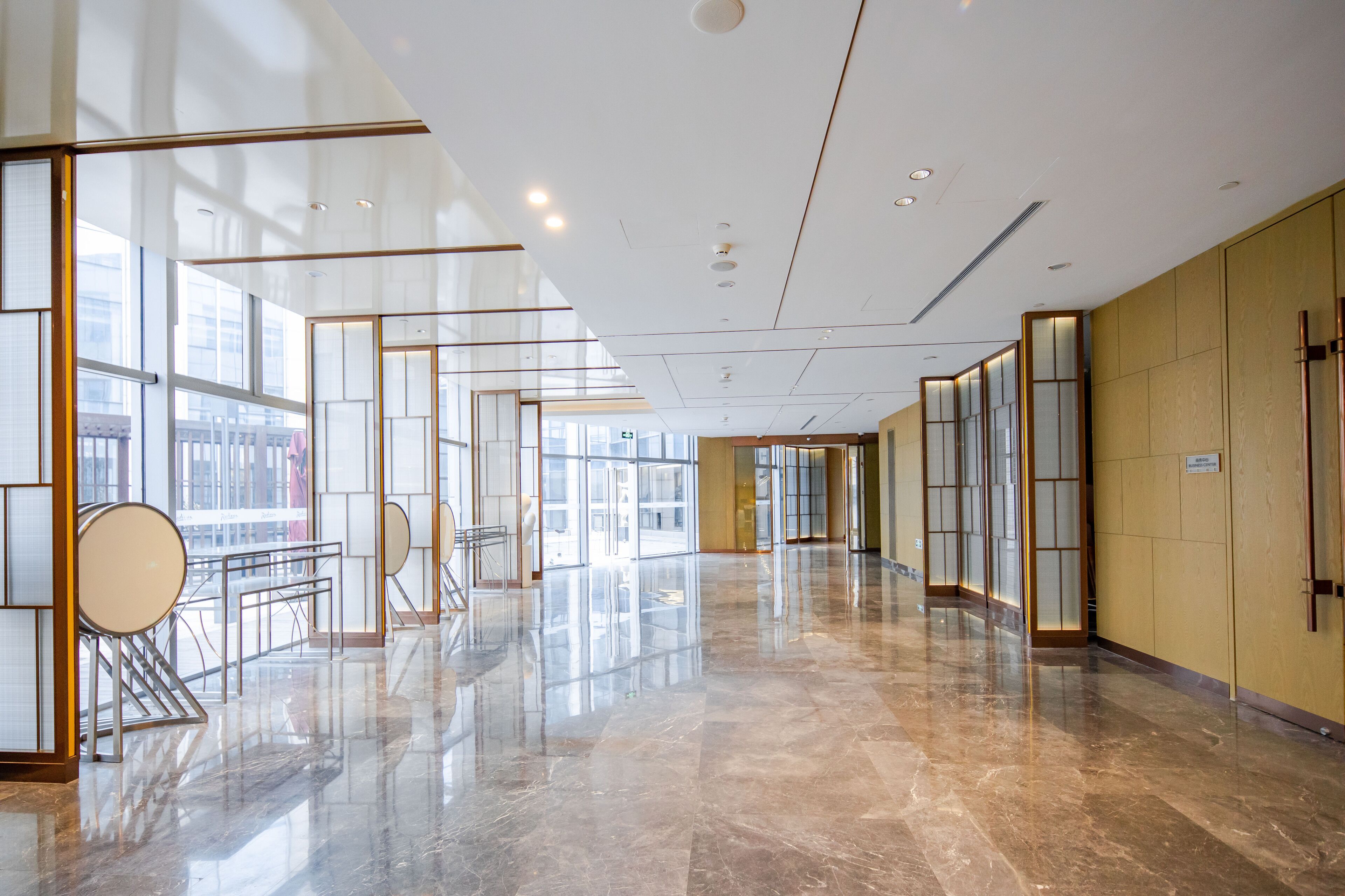 A spacious, modern hotel corridor with reflective marble floors, large windows, and contemporary furnishings.