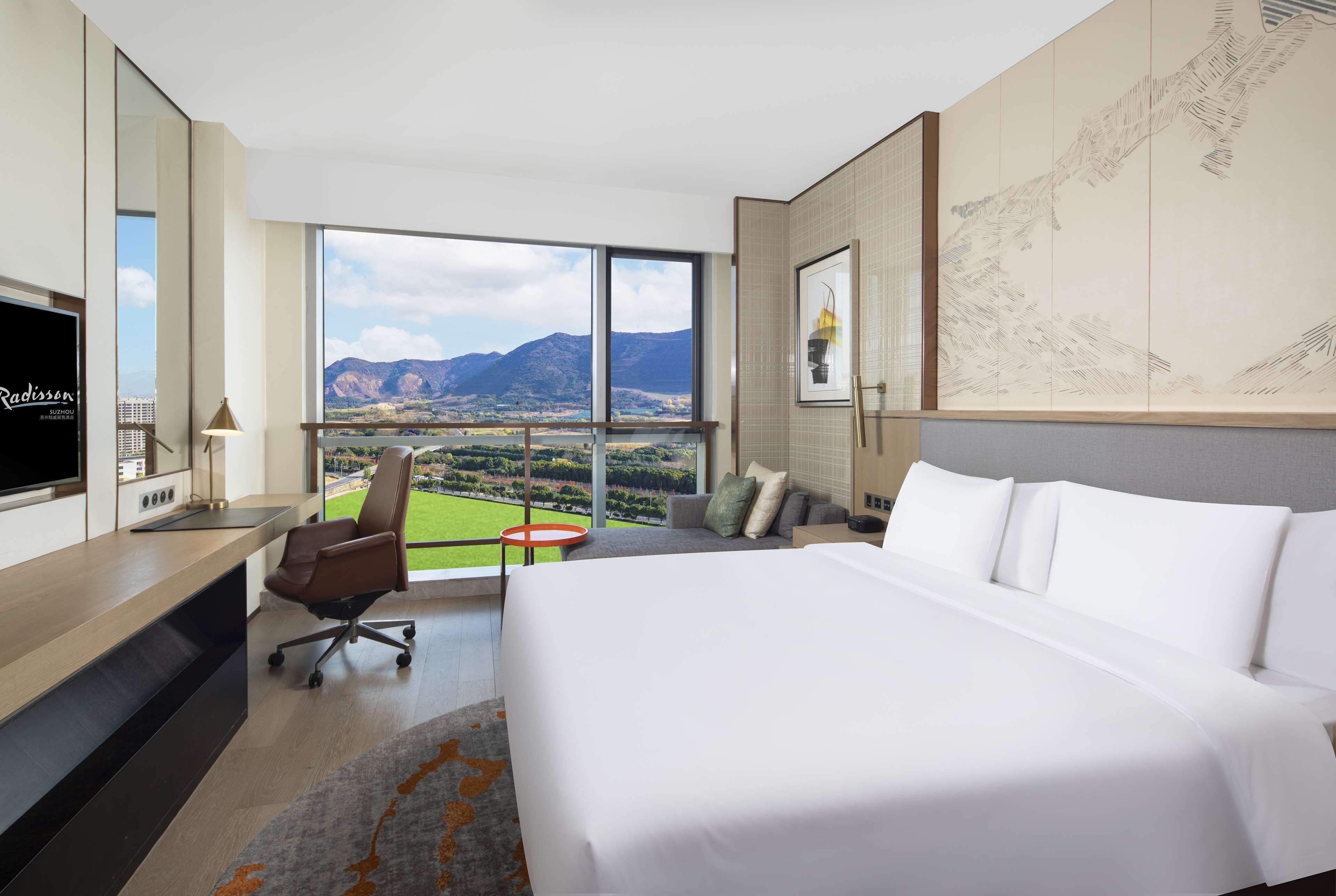 A hotel room with a large bed, a desk with a TV, a chair, and a view of mountains through large windows.