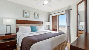 A hotel room with a large bed, two bedside tables with lamps, a ceiling fan, and a view of the sea through a sliding door. - Surf Beach 406 - Luxury Corner Unit (Treasure Island)