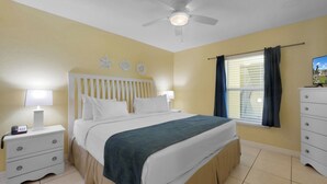 Un dormitorio con cama, mesitas de noche, cómoda, ventilador de techo y ventana con cortinas.