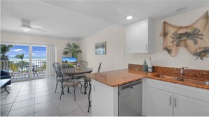 Una cocina con encimera de granito, lavavajillas y pileta. Hay un comedor con mesa y sillas, y vista a la playa a través de amplios ventanales.