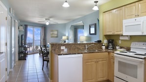 A kitchen with a dishwasher, stove, and a dining area with a chair and table. - Surf Beach 402 - Spectacular Sunsets - Beach Front (Treasure Island)