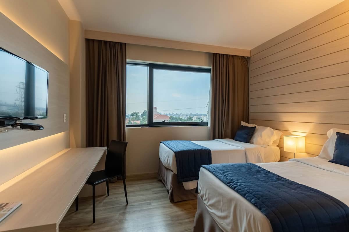 Une chambre d’hôtel avec deux lits, un bureau, une chaise et une grande fenêtre avec des rideaux.