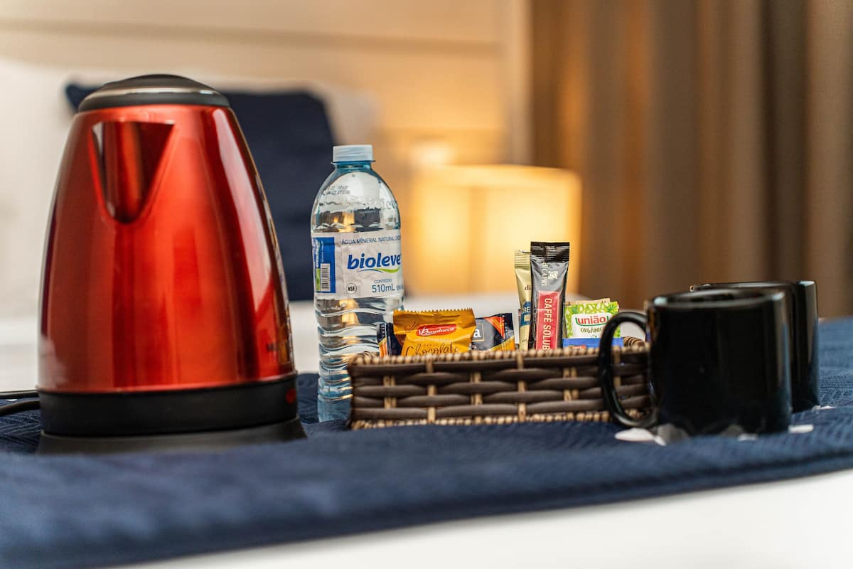 Une bouilloire rouge, une bouteille d’eau, un panier rempli d’en-cas et une tasse noire sur une table.