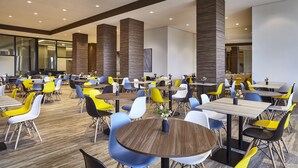 A spacious dining area with multiple tables and chairs, featuring a mix of yellow and blue chairs, and a modern interior design with wooden accents and recessed lighting. - Hotel Contemporâneo BY Royal Palm Hotels & Resorts (Campinas)