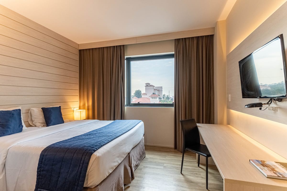 Une chambre d’hôtel avec un grand lit, un bureau, une chaise, un téléviseur fixé au mur et une fenêtre avec des rideaux.