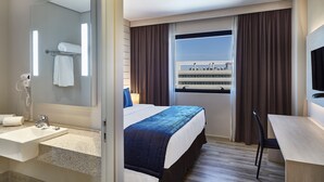A hotel room with a bed, a desk, a mirror, a sink, and a window with curtains. - Hotel Contemporâneo BY Royal Palm Hotels & Resorts (Campinas)