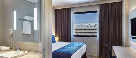 A hotel room with a bed, a desk, a mirror, a sink, and a window with curtains.