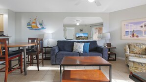 Un salon comprenant un canapé, une table basse, une table à manger et des chaises. Il y a un tableau au mur et un ventilateur de plafond.
