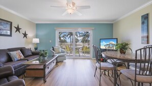 A living room with a sofa, coffee table, and TV. There is a balcony with a view of palm trees and the ocean.