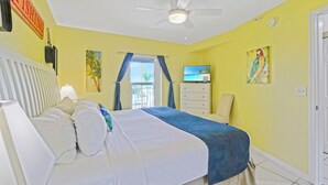 A hotel room with a bed, a ceiling fan, a TV, and a painting on the wall.