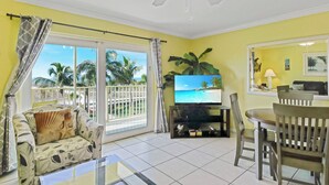 A living room with a TV, a sofa, a dining table, and a balcony with a view of palm trees.