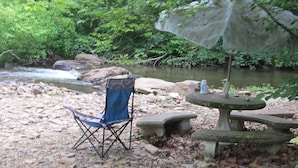 Instalación de campamento con una silla y una mesa de piedra bajo un árbol, cerca de un arroyo.
