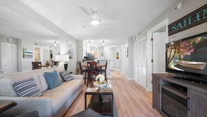 Un salon moderne avec un canapé, une table basse et un téléviseur. On y trouve également un coin repas avec des chaises et une table. Un ventilateur de plafond est visible, et les murs de la pièce sont peints en blanc, tandis que le sol est en bois.