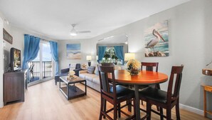 Un salon avec un coin repas, un balcon avec des rideaux bleus et un tableau représentant un pélican.