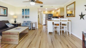 Una sala de estar con una mesa de centro de madera, un sofá y un comedor con sillas blancas. Se observa una cocina al fondo con un refrigerador y un microondas.