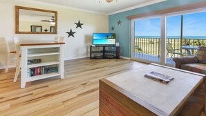 Una sala de estar con piso de madera, una estantería blanca, un televisor de pantalla plana y un balcón con vista a la playa.