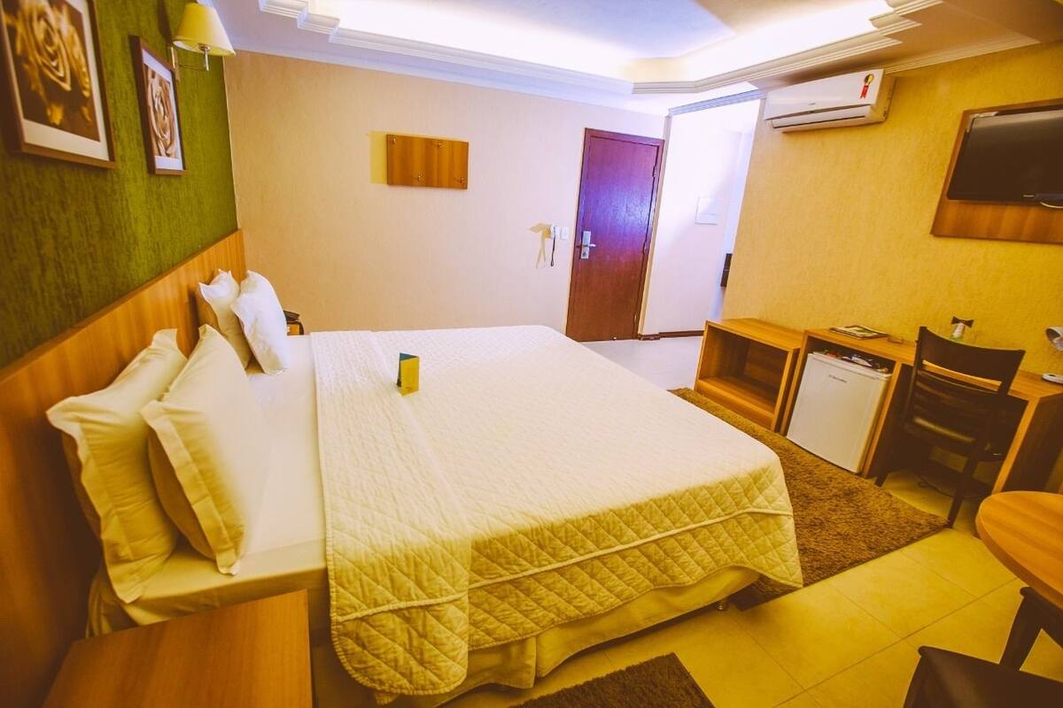 Quarto de hotel com cama, escrivaninha, cadeira e um pequeno frigorífico.