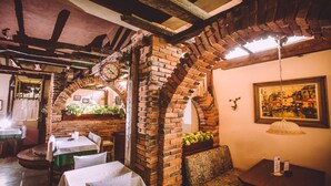 A cozy restaurant with brick arches, wooden beams, and a rustic interior. Tables are set with white tablecloths, and there are plants and a painting on the walls. - Hotel Dom Otto (Vespasiano)