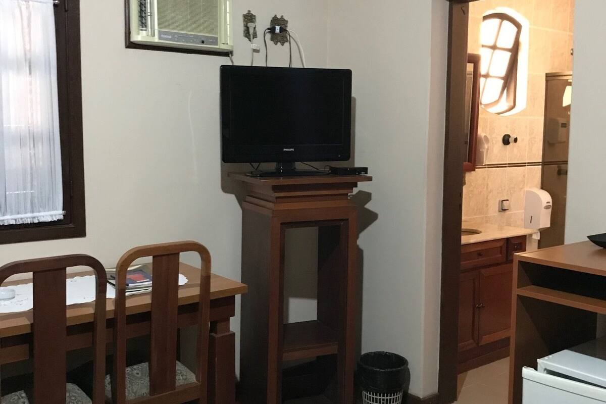 Quarto compacto com mesa de madeira, duas cadeiras, televisão, pequena geladeira e lixeira.