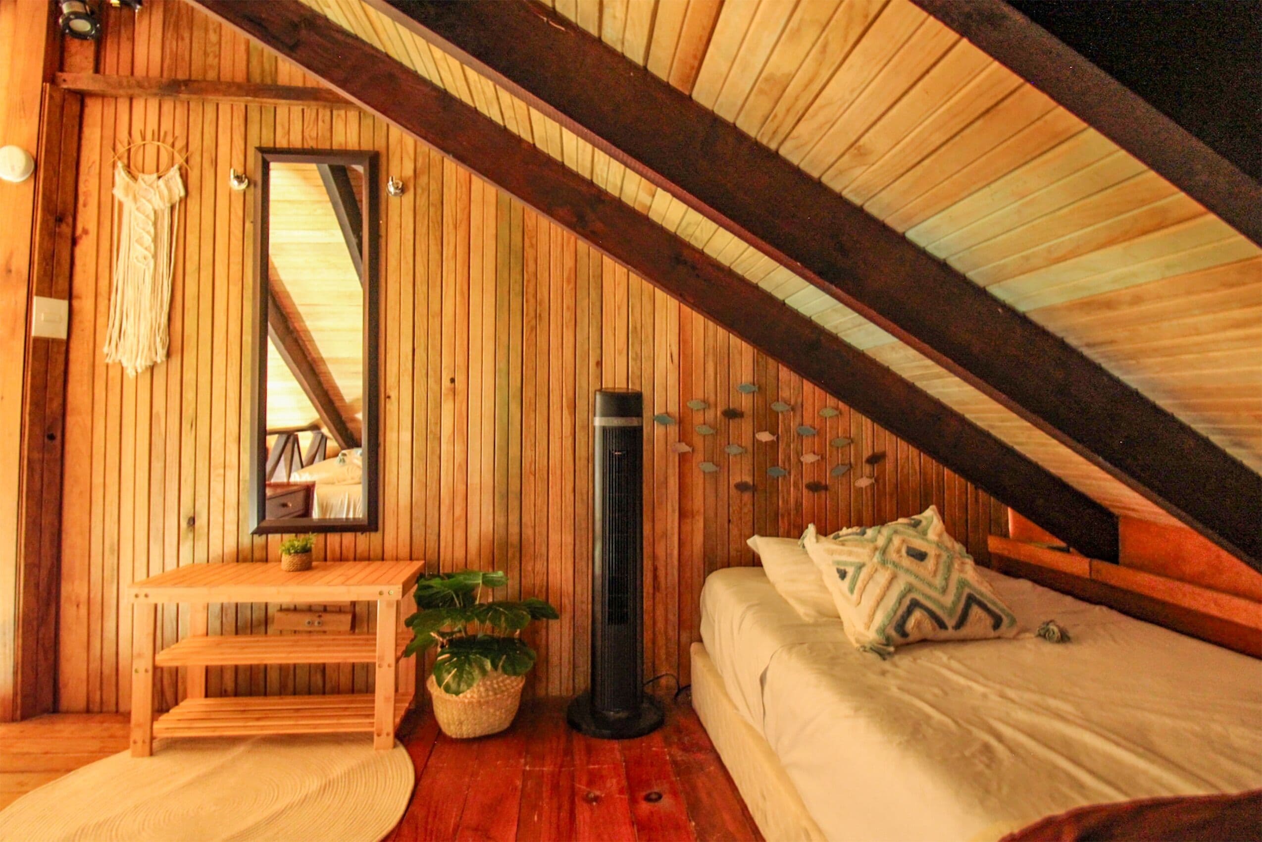 A cozy attic bedroom with wooden walls, a bed with pillows, a bedside table with a plant, a mirror, and a small bench.