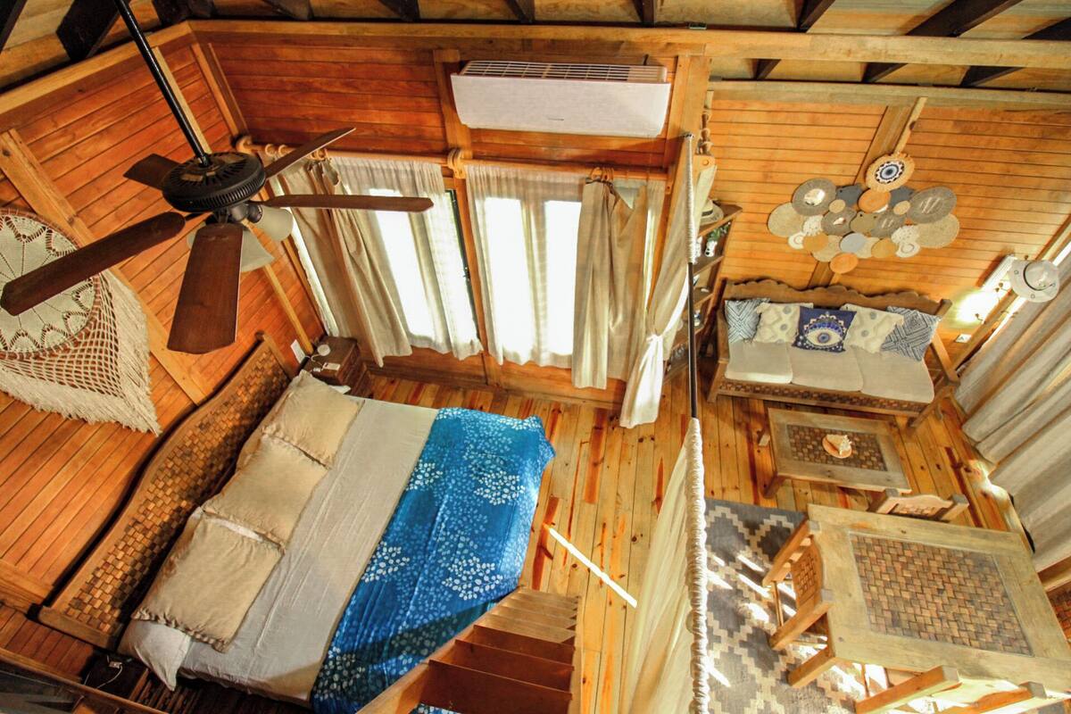 A wooden cabin with a bed, a table, a ceiling fan, and a window with curtains.