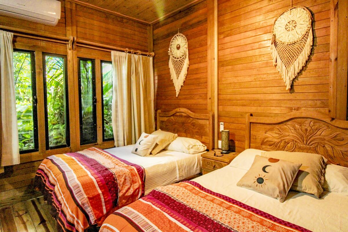 A wooden cabin room with two beds, a window with curtains, and a wall hanging.
