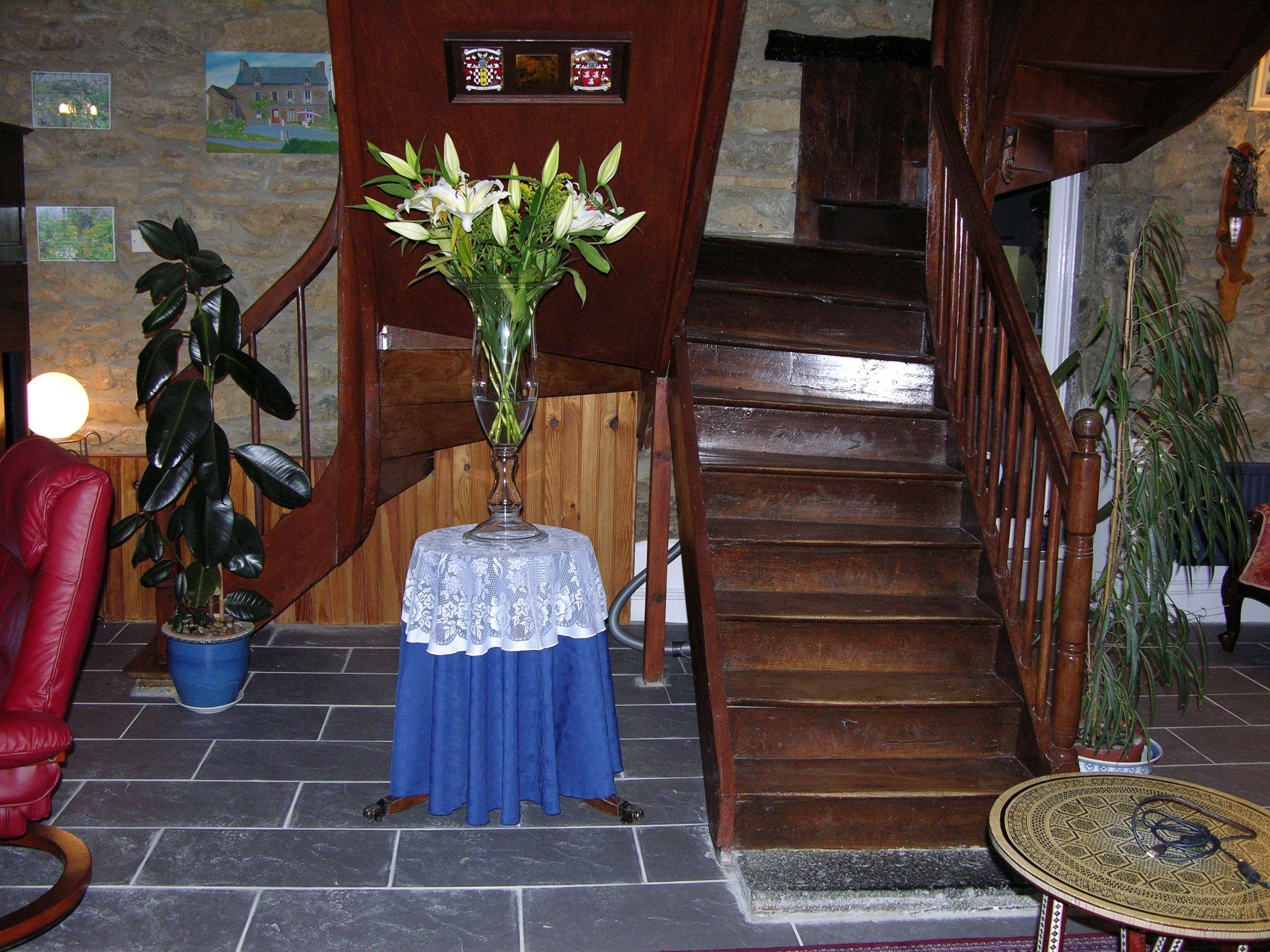 Un escalier avec une rampe en bois, une table bleue recouverte d’une nappe en dentelle et un vase rempli de fleurs.
