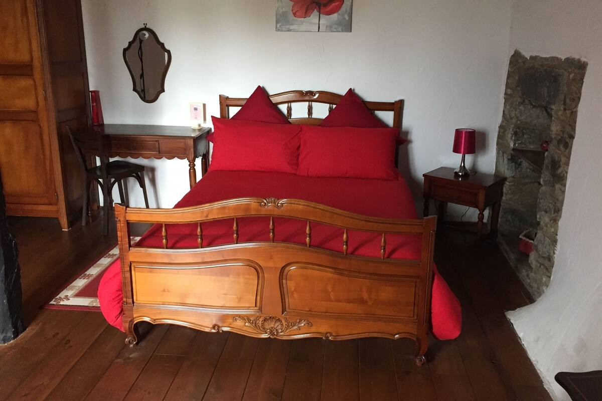 Una cama de madera con ropa de cama roja en una habitación con chimenea de piedra, un escritorio y una silla.