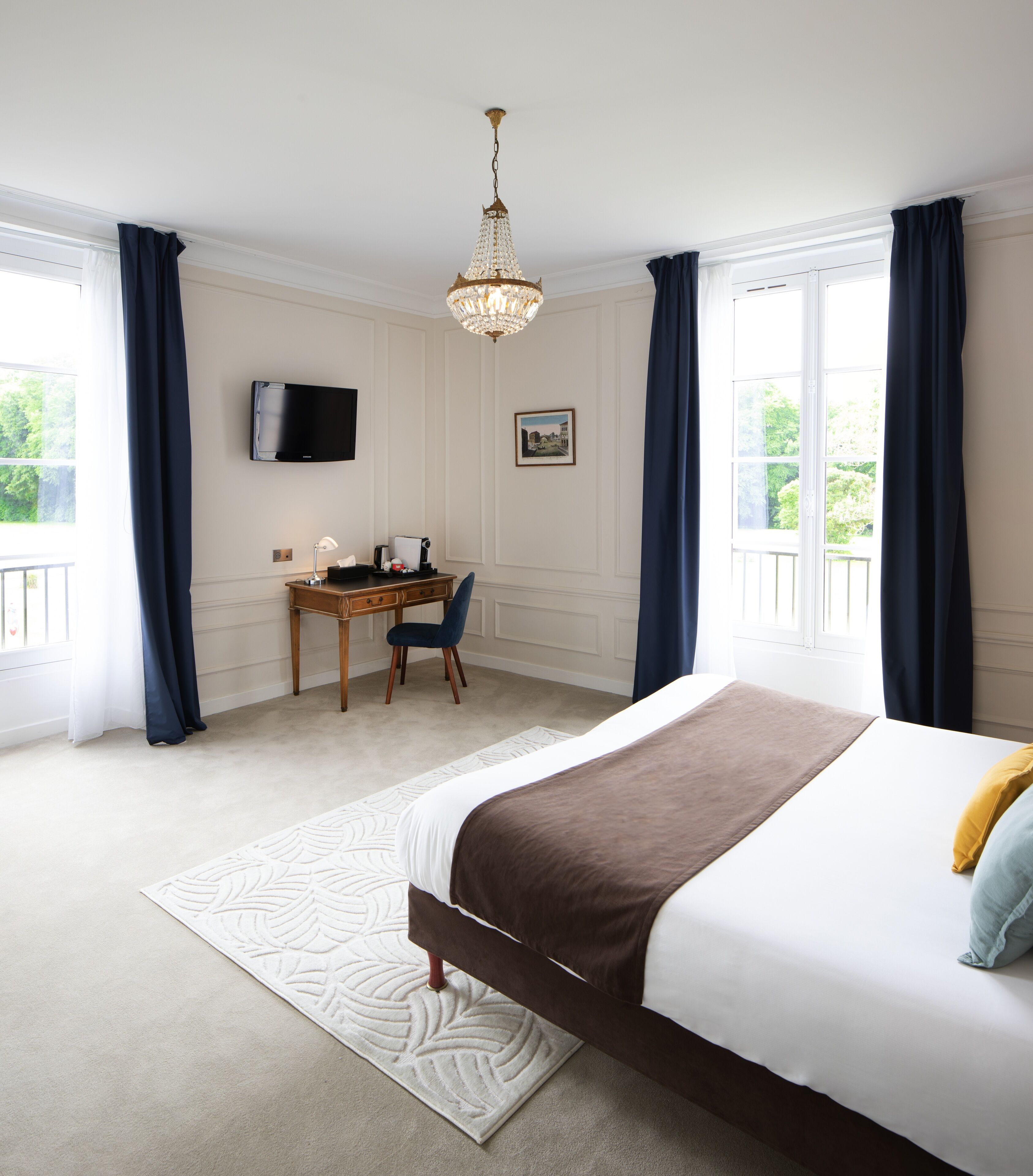 A bedroom with a bed, a desk, a chair, a television, and a chandelier.
