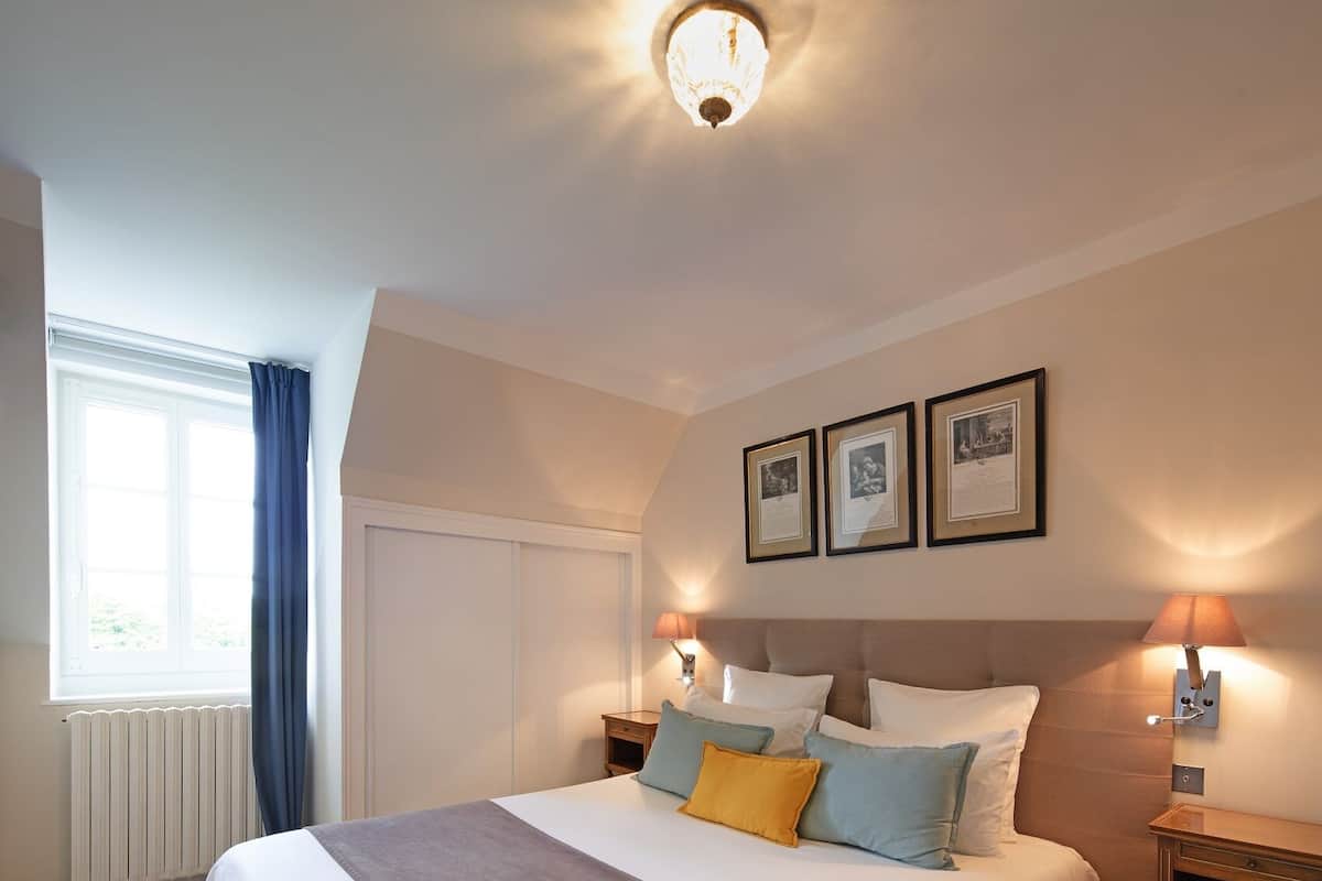 Une chambre à coucher bien rangée, avec un grand lit, des lampes de chevet, une fenêtre avec des rideaux et des tableaux encadrés accrochés au mur.