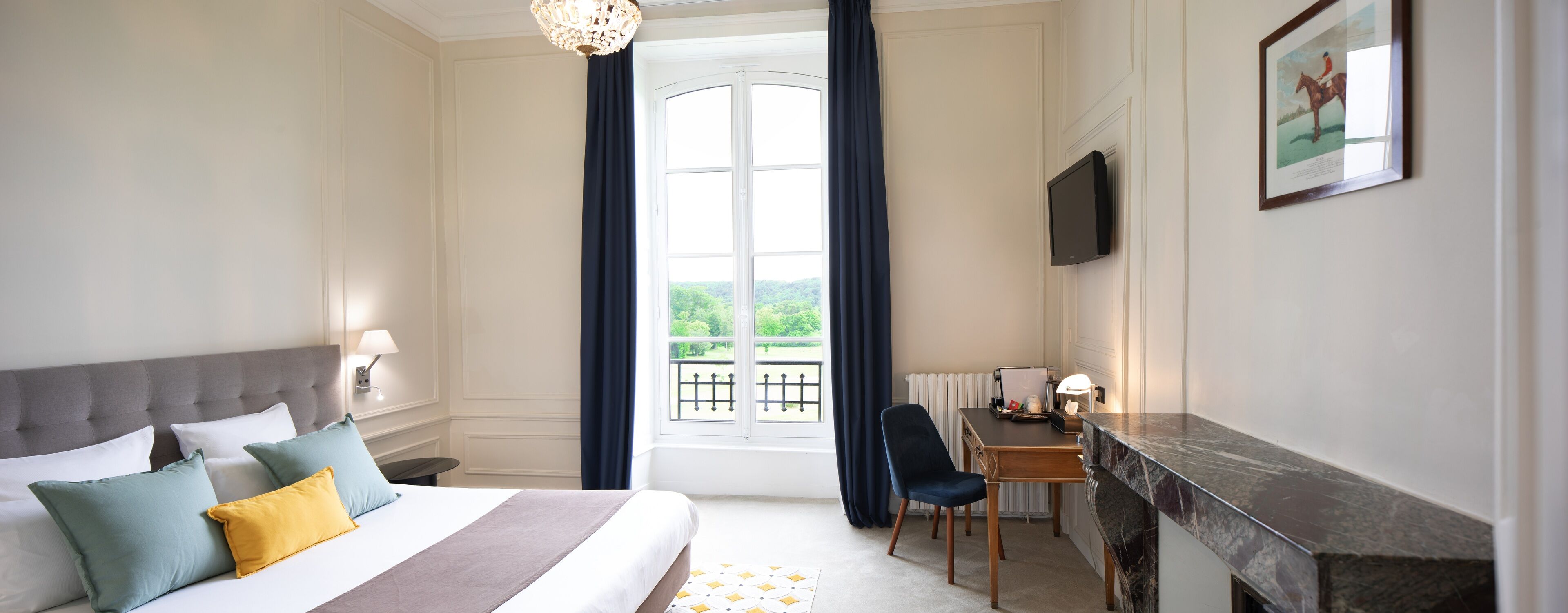 A hotel room with a bed, a desk, a chair, a TV, and a view of greenery through the window.