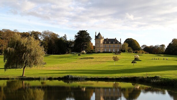 A large, historic building with a tower, surrounded by a well-maintained lawn and a pond reflecting the structure.