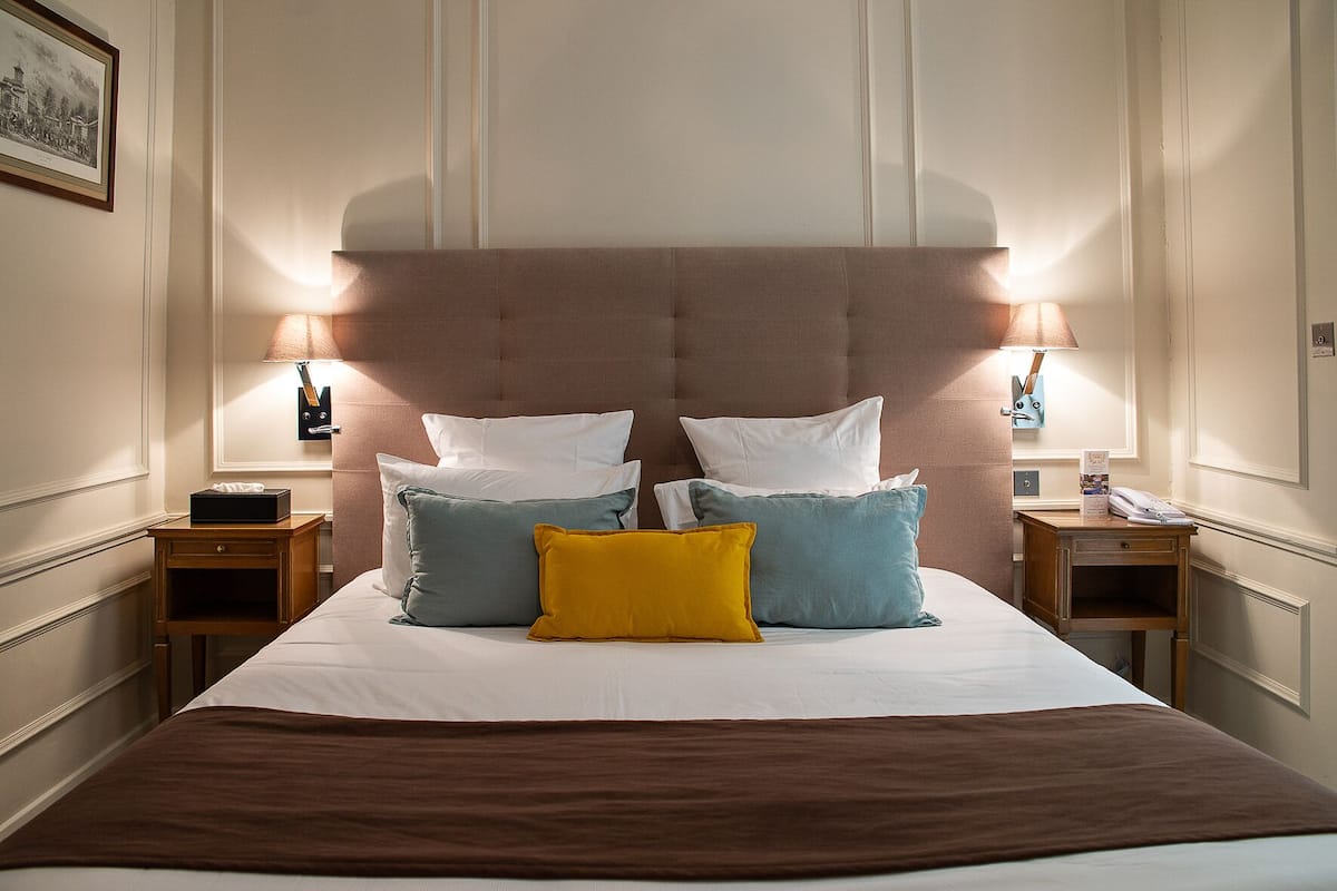 Un lit bien fait, avec une tête de lit marron, plusieurs oreillers de différentes couleurs, des tables de chevet avec des lampes et un tableau encadré au mur.