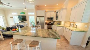A modern kitchen with white cabinets, a large island with stools, a refrigerator, microwave, and a coffee maker.