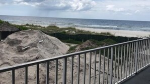A beach view from a balcony with a metal railing, sandy beach, and ocean in the background.