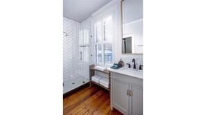 A bathroom with a herringbone tile backsplash, a freestanding shower, a window with blinds, a sink with a mirror, and wooden flooring. - 154 ABC Spring Street - 7 BR / 5 BA Holiday home 7 (Charleston)