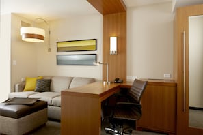 A hotel room with a wooden desk, a leather chair, a sofa with cushions, a lamp, and two framed artworks on the wall.