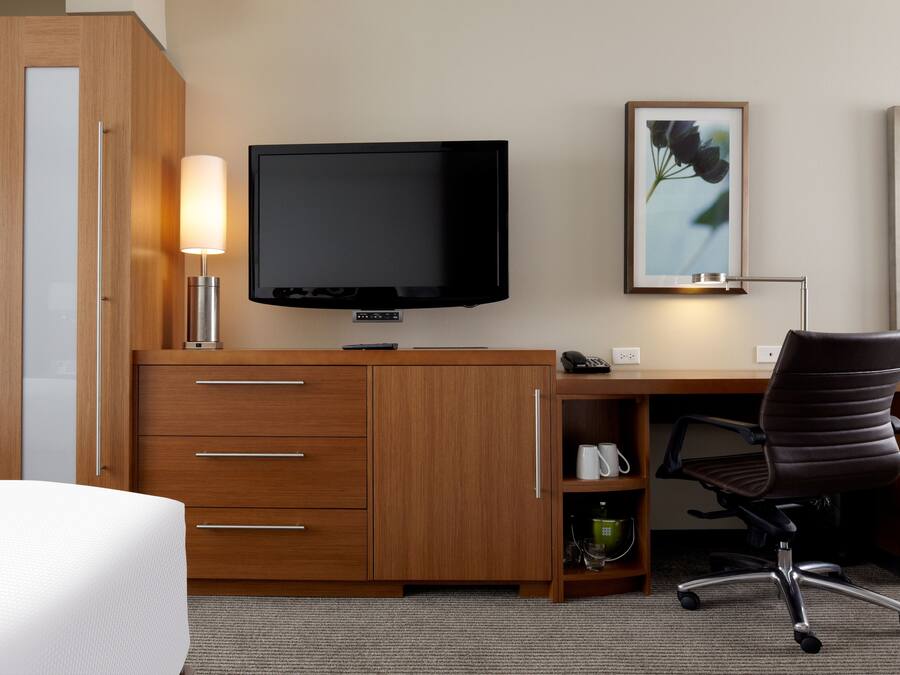 A hotel room with a wooden desk, a flat-screen TV, a lamp, a chair, and a framed picture on the wall.