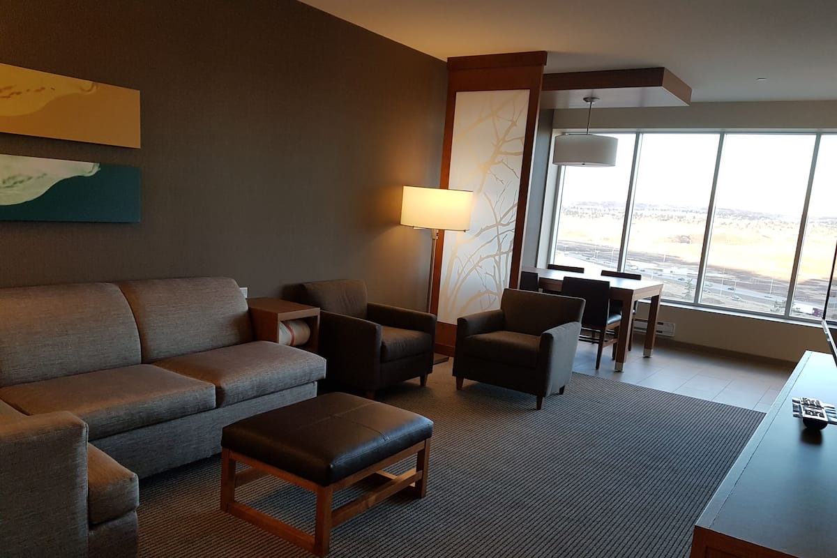 A modern hotel room with a sofa, armchairs, a small table, and a lamp. There is a large window offering a view of the city.