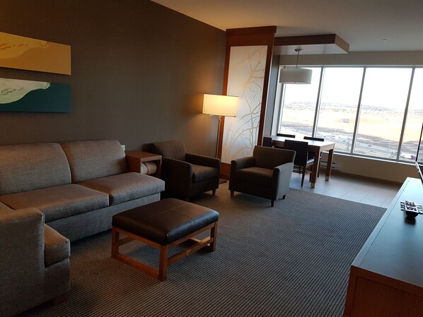 Una habitación de hotel moderna con sofá, sillones, una mesita y una lámpara. Cuenta con un ventanal que ofrece vistas a la ciudad.