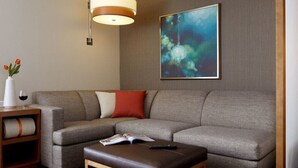 Una sala de estar moderna con un sofá gris, una mesita de madera con un florero y una copa de vino, un reposapiés marrón y un cuadro abstracto grande en la pared.