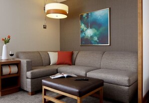 A modern living room with a grey sofa, a wooden side table with a vase and wine glass, a brown ottoman, and a large abstract painting on the wall. - Hyatt Place Calgary Airport (Calgary)