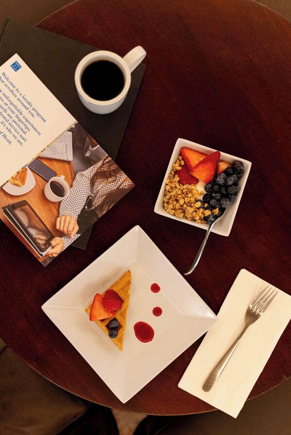 Uma mesa com uma xícara de café, uma tigela de granola com frutas, uma fatia de bolo com morangos e mirtilos, um cardápio e um garfo sobre um guardanapo.