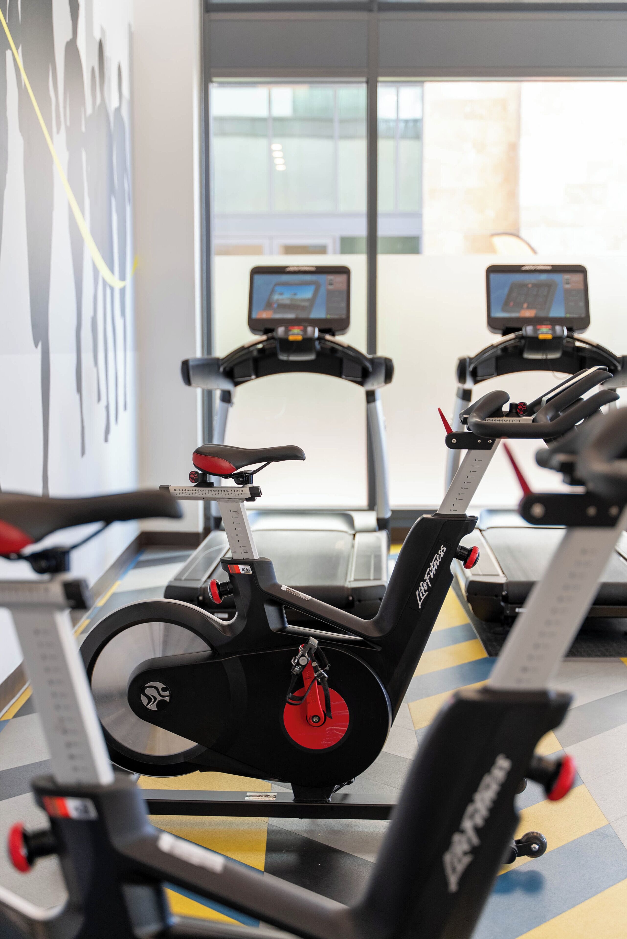 A gym with stationary bikes and treadmills, a large window, and a wall with abstract art.