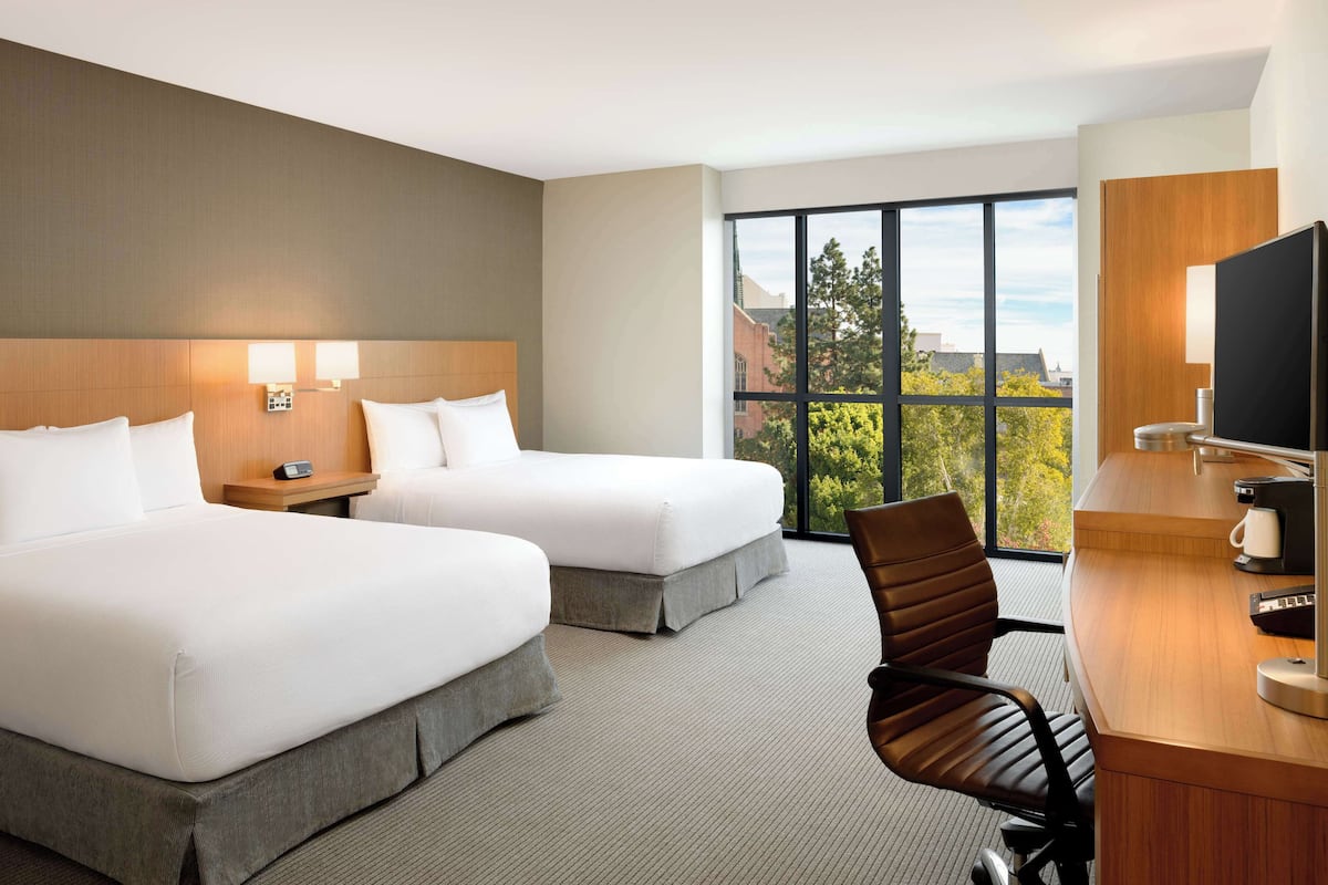A hotel room with two beds, a desk, a chair, a TV, and a large window with a city view.